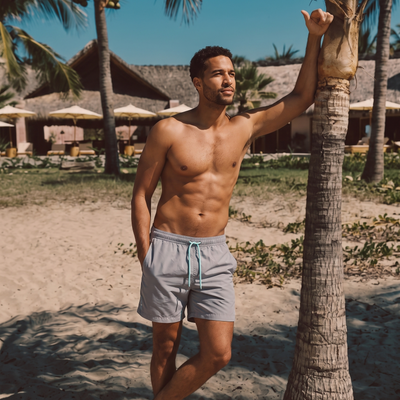 A shirtless man standing on a sandy beach next to a palm tree, wearing Misty Grey Blauw Short quick-dry swim trunks