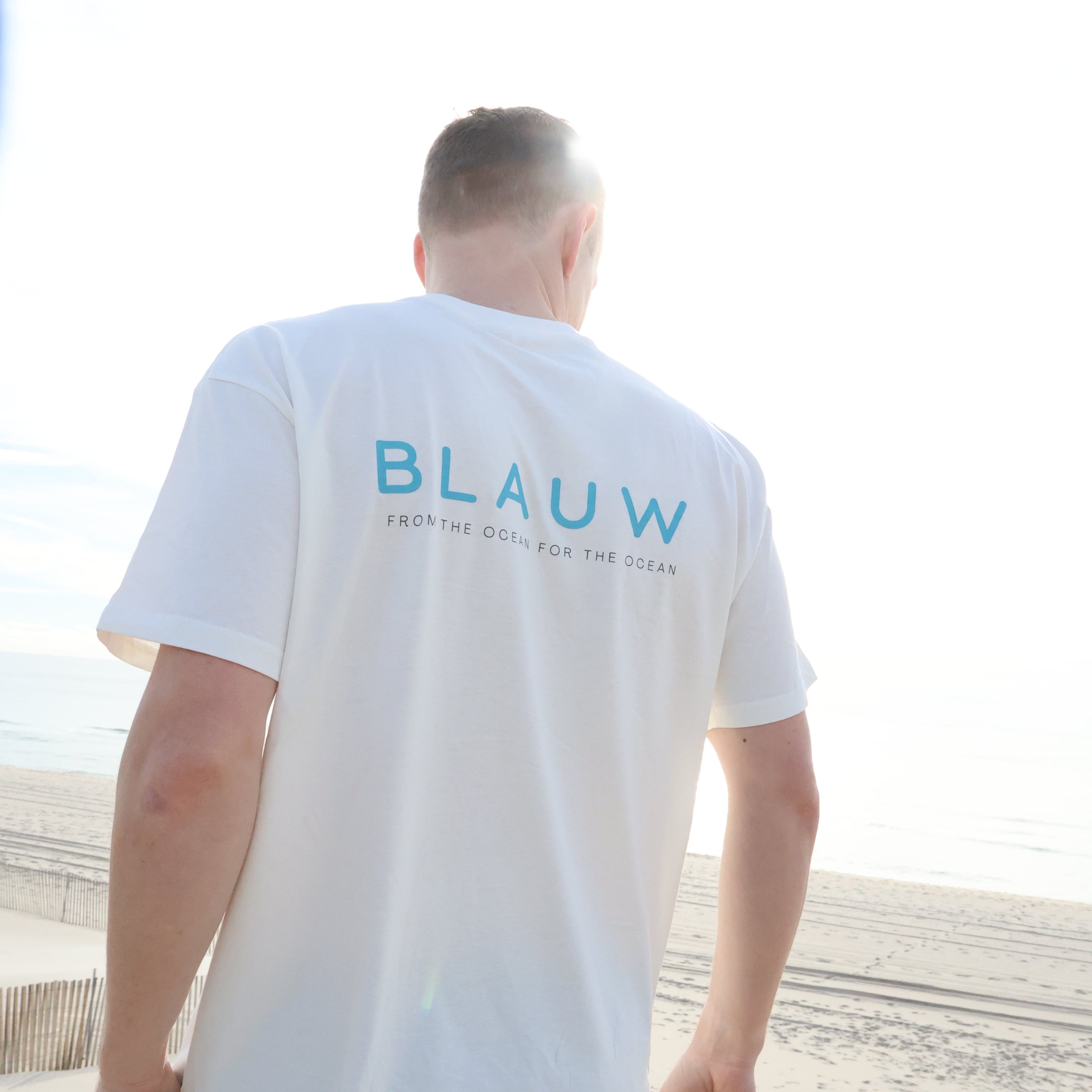 A man wearing a white organic cotton t-shirt with 'BLAUW' printed in blue on the back, standing on a sandy beach.