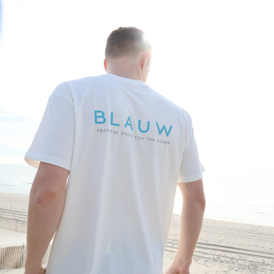 A man wearing a white organic cotton t-shirt with 'BLAUW' printed in blue on the back, standing on a sandy beach.