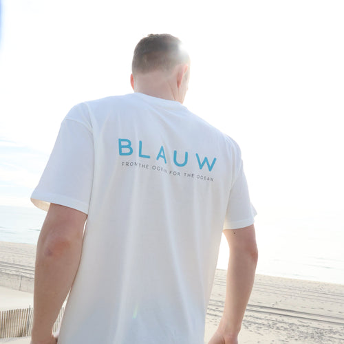 A man wearing a white organic cotton t-shirt with 'BLAUW' printed in blue on the back, standing on a sandy beach.