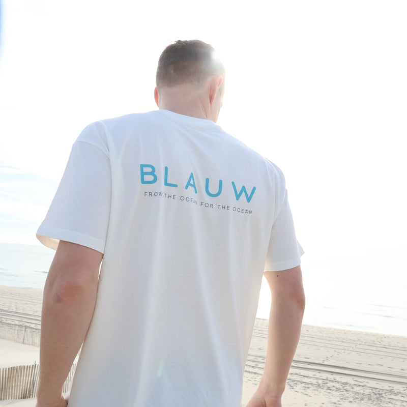 A man wearing a white organic cotton t-shirt with 'BLAUW' printed in blue on the back, standing on a sandy beach.