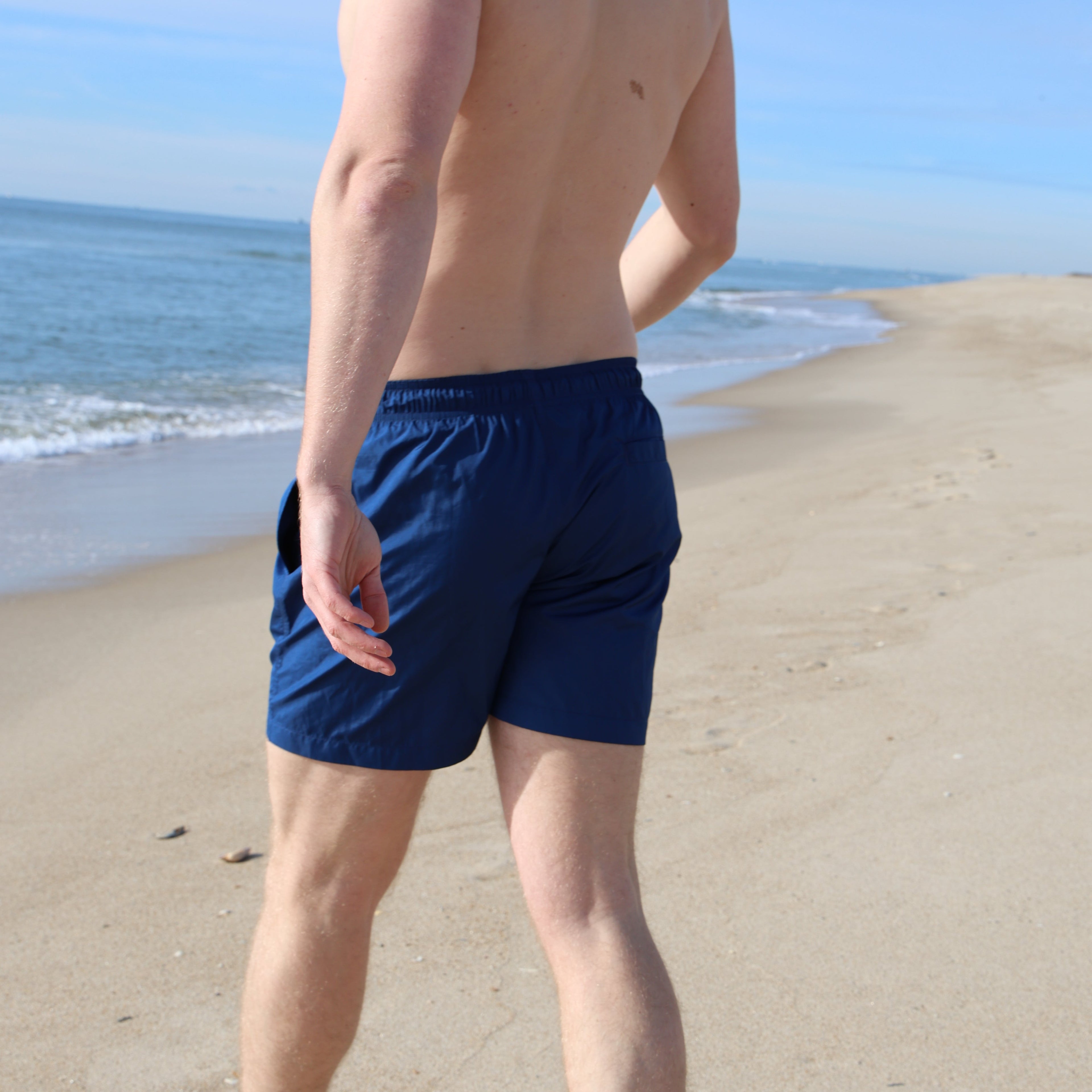 Person wearing blue sustainable swimwear for men, standing on a sandy surface with the blue ocean in the background.