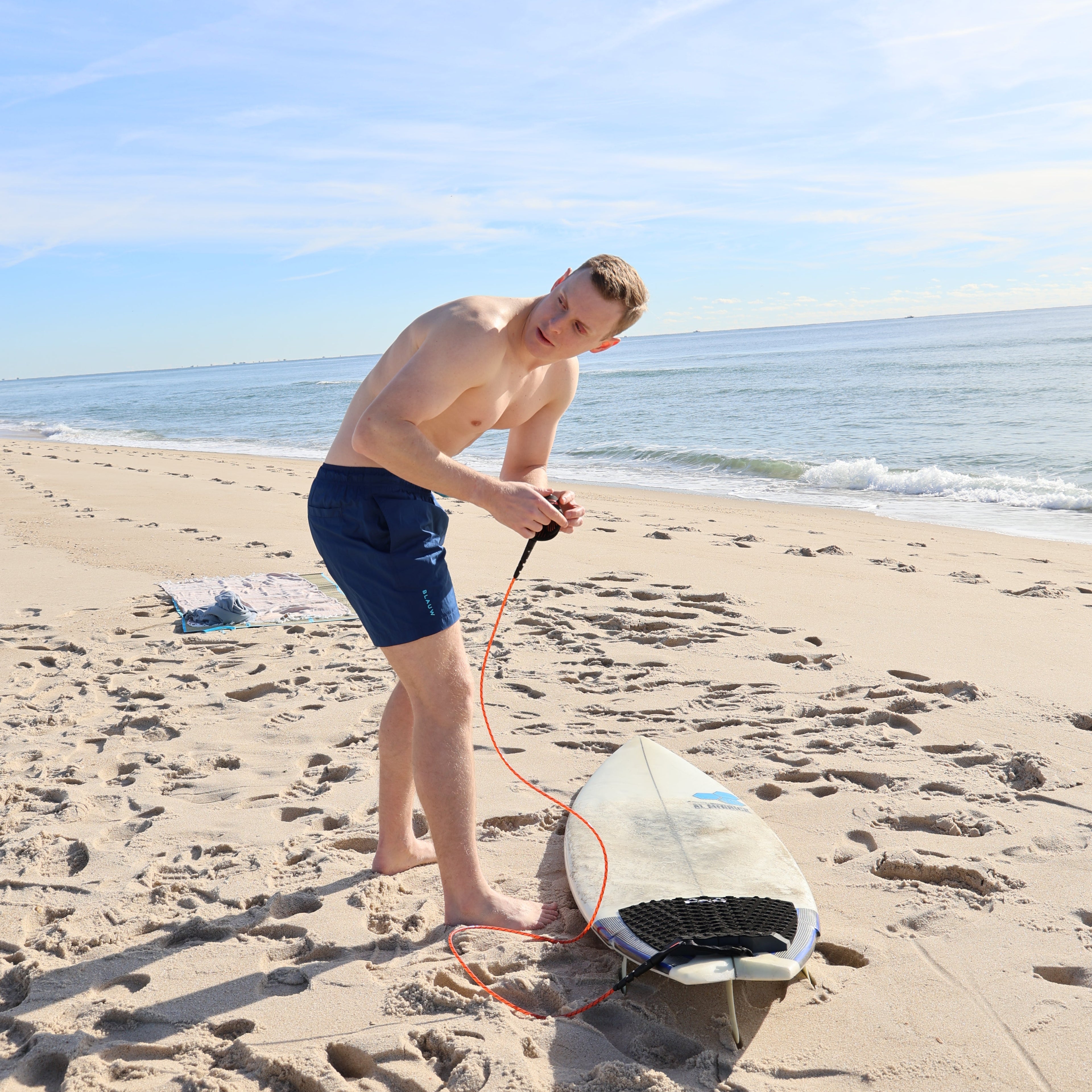 Person on a beach with a surfboard under a clear blue sky, wearing sustainable swimwear from ocean plastic.