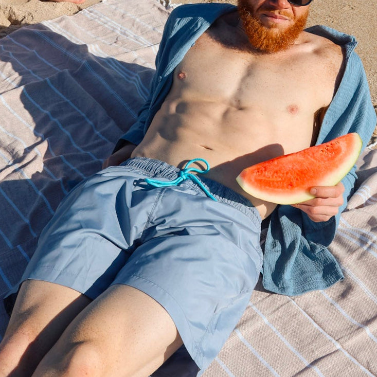 Person laying down on a sandy beach, holding watermelon slices and wearing sustainable swimwear.