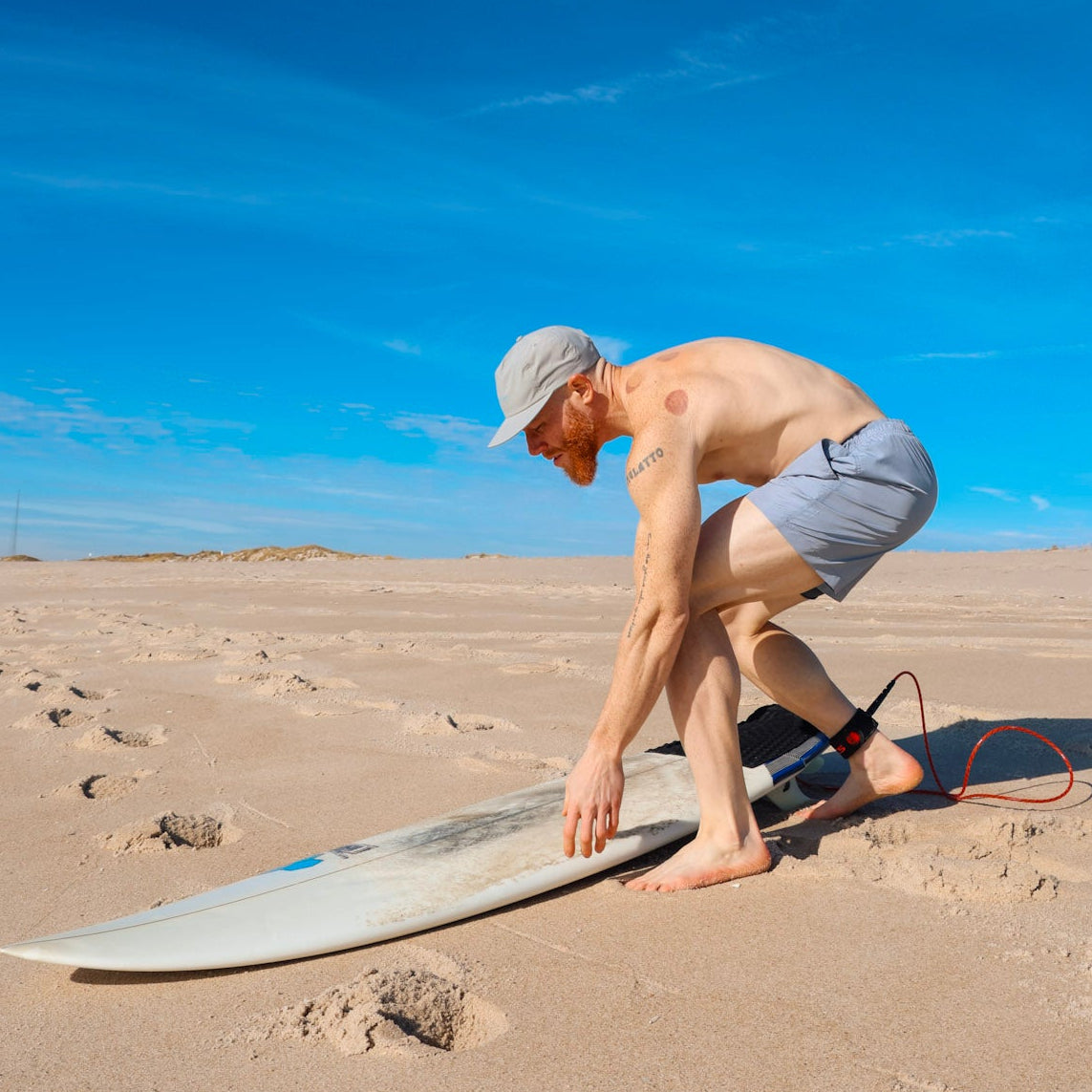 Man with a surfboard on a sandy beach wearing recycled swim shorts