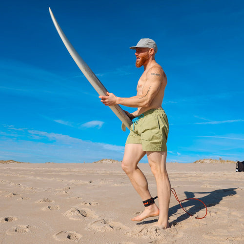 Man holding a surfboard on a sandy beach wearing a comfortable swim short from ocean plastic.