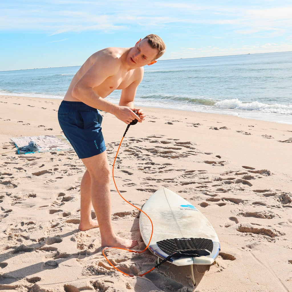 Person on a beach with a surfboard under a clear blue sky, wearing sustainable swim shorts.