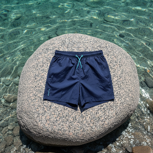 Navy blue sustainable swim shorts, made from recycled materials, on a stone by clear blue water