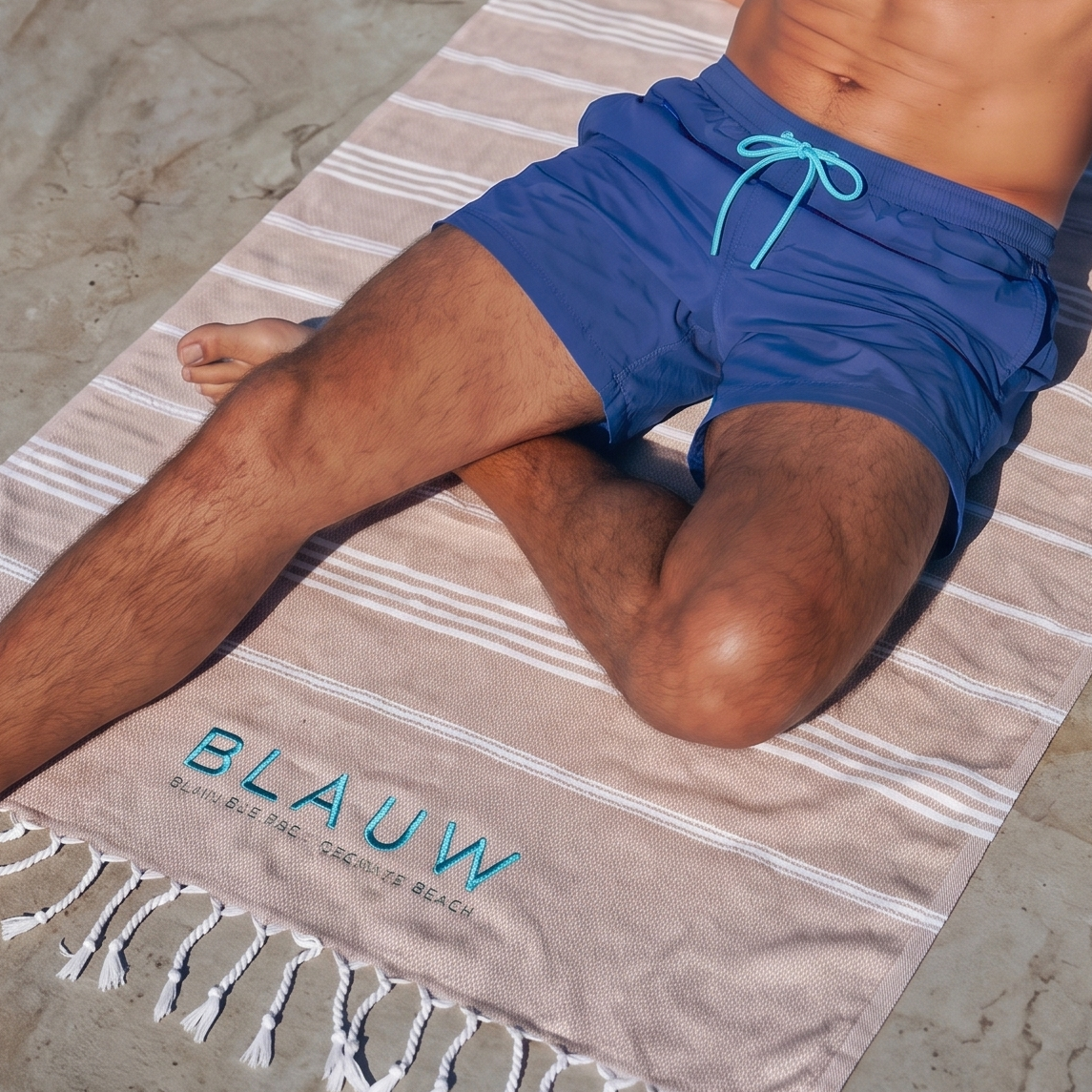 A man relaxes on a striped beach towel with white fringe, positioned on a stone dock with the blue ocean water in the background. The towel features the brand name 'BLAUW' printed in light blue near the bottom.