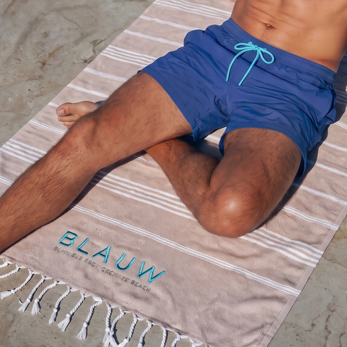 A man relaxes on a striped beach towel with white fringe, positioned on a stone dock with the blue ocean water in the background. The towel features the brand name 'BLAUW' printed in light blue near the bottom.