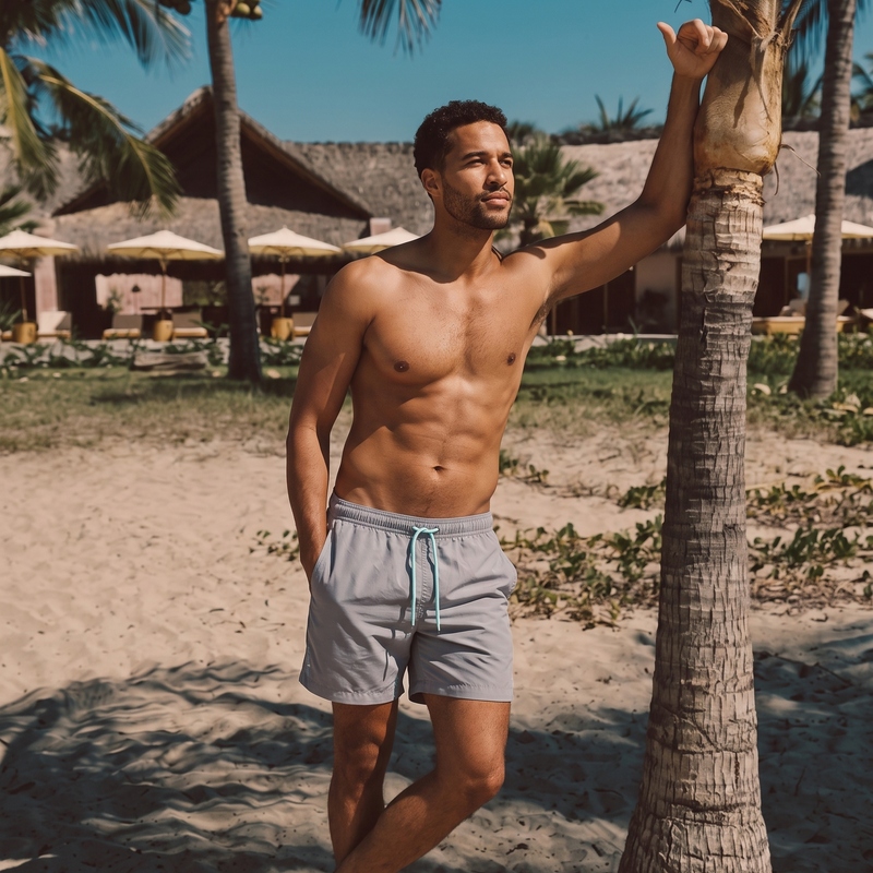 A shirtless man standing on a sandy beach next to a palm tree, wearing Misty Grey Blauw Short quick-dry swim trunks