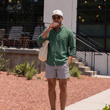 A man wearing a green button-down shirt, Misty Grey Blauw Short, a white cap, and sunglasses stands outdoors on a sidewalk in front of a modern building, holding a coffee cup and carrying a cream tote bag.